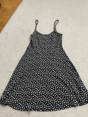 Svart blommig sommarklänning - Söt svart klänning med vita blommor och tunna axelband. Klänningen har en lätt utställd passform och är perfekt för varma dagar. Materialet känns mjukt och luftigt, vilket gör den skön att bära hela dagen.