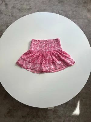 Rosa kortkjol från Gina Tricot - Supersöt rosa kortkjol från Gina Tricot med smock i midjan och volangkant💓. Kjolen har ett blommigt mönster och är gjord i ett lätt och luftigt material, perfekt för varma dagar. Den är i storlek 158/164 och nästan helt oanvänd🌷