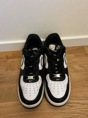 Nike Air Force 1 svartvita sneakers - Säljer ett par klassiska Nike Air Force 1 sneakers i svart och vitt läder. Skorna har svart sula, vita detaljer och den ikoniska swoosh-loggan på sidan. Låga med svart snörning och perforerad tå för extra ventilation. Perfekta för en clean och stilren look.