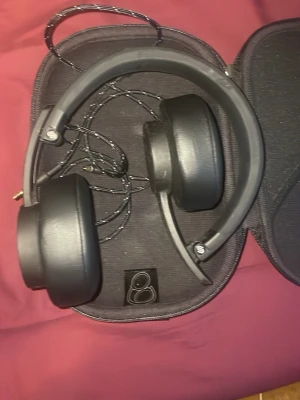 Svarta over-ear hörlurar med sladd - Säljer ett par svarta over-ear hörlurar med vadderade kåpor och justerbart huvudband. Hörlurarna har en sladd med tygklädd kabel och kommer med ett hårt fodral för enkel förvaring. Perfekta för dig som vill ha bra ljud och komfort under längre lyssningssessioner.