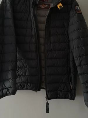 Svart pufferjacka från Parajumpers - Snygg svart pufferjacka från Parajumpers med gul detalj vid halsen och patch på ärmen. Jackan har quiltad design, dragkedja framtill och sidofickor med dragkedja. Perfekt för kalla dagar och ger en sportig look. Har även en lagning på ena armen