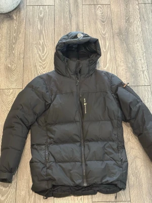 Svart dunjacka från Sail Racing - Snygg svart dunjacka från Sail Racing med huva och dragkedja framtill. Jackan har två sidofickor med dragkedja och en bröstficka med logga. Perfekt för kalla vinterdagar tack vare det tjocka dunfodret och den höga kragen.