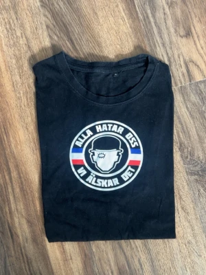 Svart t-shirt med tryck - Svart t-shirt med rund hals och tryck framtill som säger 'Alla hatar oss vi älskar det' i vitt, blått och rött. 