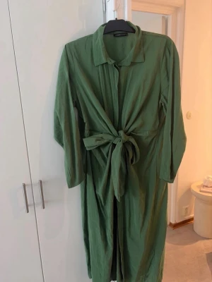 Grön långklänning med knytband - Stilren grön långklänning med krage, långa ärmar och ett stort knytband i midjan. Klänningen har en avslappnad passform och knappar framtill. Perfekt för dig som gillar enkel men snygg design. Använd en gång. Liten k storlek passar mig som har L/XL
