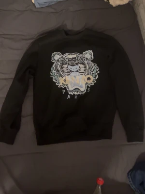 Svart Kenzo sweatshirt med tigertryck - Svart sweatshirt från Kenzo med ikoniskt tigertryck och broderad logga på bröstet. Tröjan har rund halsringning, långa ärmar och ribbade muddar. Perfekt för dig som vill ha en statement piece i garderoben.