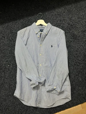 Blårandig skjorta från Ralph Lauren - Klassisk blårandig skjorta från Ralph Lauren med vit knappstängning och broderad logga på bröstet. Skjortan har lång ärm, krage och är tillverkad i mjuk bomull. Perfekt för en stilren och avslappnad look.