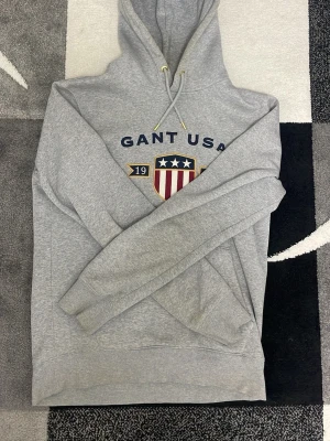 Grå hoodie från Gant USA - Snygg grå hoodie från Gant USA med tryckt logga och sköld på bröstet. Klassisk huva med dragsko och ribbade muddar vid ärmslut och nederkant. Tillverkad i mjukt sweatshirtmaterial som är skönt att ha på sig. Perfekt för en avslappnad och sportig stil. Priset kan diskuteras.