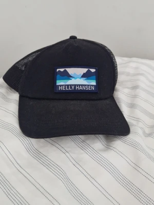 Svart keps från Helly Hansen - Säljer en svart keps från Helly Hansen med mesh på sidorna och bak. Framtill sitter en patch med bergsmotiv i blå toner och texten 'Helly Hansen'. Klassisk böjd skärm och justerbar passform bak.