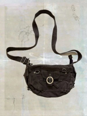 Brun axelväska med detaljer - Cool brun axelväska i canvas med justerbar rem och flera fickor. Väskan har silverfärgade metalldetaljer, ringar och ett spänne framtill. Perfekt för dig som gillar en avslappnad och funktionell stil.