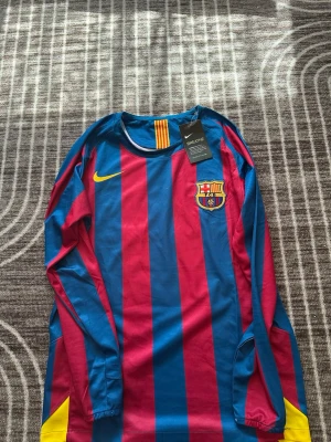 Barcelona långärmad matchtröja Nike - Snygg långärmad Barcelona matchtröja från Nike med klassiska blå och röda ränder. Tröjan har klubbmärke på bröstet, gult Nike-märke och Ronaldinho 10 tryckt på ryggen. Tillverkad i lätt och ventilerande material, perfekt för fotboll eller streetwear.
