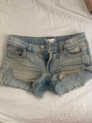Ljusblå slitna jeansshorts - Säljer ett par ljusblå jeansshorts med råa kanter och slitningar framtill. Shortsen har fem fickor, knappgylf och snygga broderade detaljer på bakfickorna. Perfekta för sommardagar och festivaler.