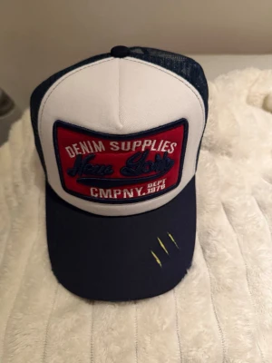 Blå och vit truckerkeps New Lady - Snygg truckerkeps från New Lady med vit front, blått nät bak och böjd blå skärm. Stor broderad patch framtill med texten 'Denim Supplies' och gula detaljer på skärmen. Perfekt för en avslappnad streetwear-look.