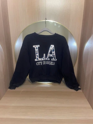 Svart sweatshirt LA City of Angels - Cool svart sweatshirt med stort LA-tryck i leopardmönster och texten 'City of Angels' framtill. Tröjan har rund halsringning och ribbade muddar vid ärmar och nederkant och är i storlek 146-152. Perfekt för en avslappnad streetstyle-look.skriv för fler bilder, pris kan diskuteras!💗