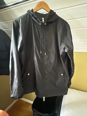 Moncler windbreaker - Snygg svart vindjacka från Moncler med huva och dragkedja framtill. Jackan har två fickor med tryckknappar och Moncler-logga på ärmen. Justerbar huva med snörning och diskreta detaljer i vitt och rött på huvan. Perfekt för blåsiga dagar.