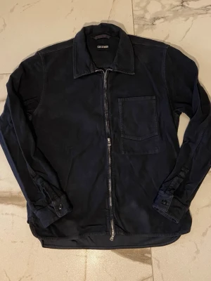 Svart overshirt/Jacka från C.O.F. Studio-S - Stilren svart overshirt/jacka från C.O.F. Studio med rak passform, bröstficka och dragkedja framtill. Tillverkad i ett mjukt bomullstyg och har klassisk krage samt knappslå vid ärmsluten. Perfekt lager-på-lager-plagg för en avslappnad look. Storlek S. Made in Italy.