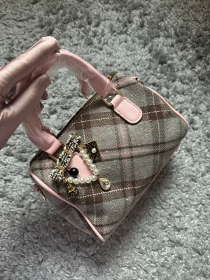 Rutig handväska med pärldetaljer - Söt handväska i rutigt tyg med rosa detaljer och handtag i rosa skinnimitation. Väskan har en dekorativ brosch med pärlor och strass framtill samt dragkedja upptill. Perfekt accessoar för en trendig look.