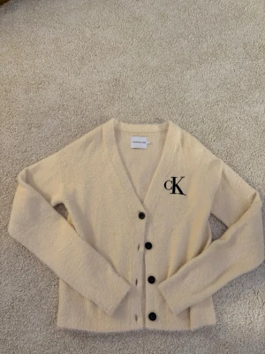 Beige stickad kofta från Calvin Klein Jeans - Mysig beige kofta från Calvin Klein Jeans med fluffig textur och svart CK-logga på bröstet. Koftan har v-ringning, långa ärmar och stängs med svarta knappar framtill. Perfekt för lager på lager och enkel att styla till olika outfits. Jag är osäker på vad storlek 16 betyder. Tröjan är använd ett mycket fåtal gånger och är i mycket bra skick (använt ca 2-3 gånger). Skriv gärna om ni ha några frågor 😊