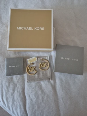 Guldiga MK örhängen från Michael Kors - Snygga örhängen i guldfärgad metall från Michael Kors med stora MK-loggor i cirkelformad design. Kommer med original ask och påse, perfekt för dig som gillar statement-smycken och exklusiv känsla.