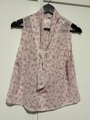 Blommig ärmlös blus med knytband - Söt ljusrosa blus med småblommigt mönster i lila och vitt. Blusen är ärmlös och har ett dekorativt knytband vid halsen. Tillverkad i ett tunt, lätt material som ger ett luftigt intryck. Perfekt för en trendig och feminin look.