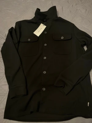 Svart overshirt från Only & Sons - Stilren svart overshirt från Only & Sons med knäppning framtill och två bröstfickor med lock och knapp. Overshirten har hög krage och är tillverkad i ett mjukt, tjockare tyg som ger en schysst look. Perfekt att bära öppen eller stängd över en t-shirt.