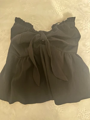 Svart linnetopp med knyt fram - Supersnygg svart linnetopp med breda axelband och stor knytning framtill. Toppen har en lätt rynkad effekt upptill och nedtill för en söt och trendig look. Perfekt att styla med jeans eller kjol för en cool outfit.