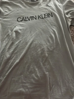 Ljus grå Calvin Klein t-shirt - Snygg ljus grå t-shirt från Calvin Klein Jeans med svart logotyptryck över bröstet. Klassisk passform och rund halsringning. Tillverkad i mjuk bomull som känns skön mot huden. Perfekt till jeans eller shorts för en clean look. Storlek 14 = M 