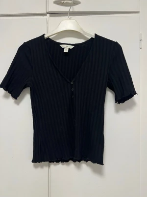 Svart ribbad topp från H&M - Svart ribbad topp från H&M med v-ringning och knappar framtill. Toppen har korta ärmar med vågig kant och är tillverkad i mjuk bomullsmix. Perfekt till jeans eller kjol för en enkel och snygg look.