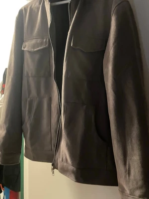 Grå mockajacka med dragkedja - Snygg grå mockajacka med raka linjer och fyra fickor framtill. Jackan har hel dragkedja och klassisk krage. Perfekt för dig som gillar stilren och enkel design.