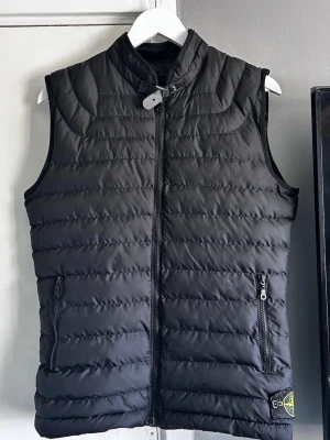 Svart dunväst från Stone Island - Snygg svart dunväst från Stone Island med quiltad design, hög krage och dragkedja framtill. Västen har två fickor med dragkedja och en ikonisk Stone Island-patch på nederkanten. Perfekt för lager-på-lager och streetwear-stil.