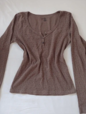 Söt brun långärmad topp med knappar - Brun långärmad topp med små diskreta mönster och tre knappar vid halsringningen. Toppen har en rundad urringning och är i mjukt bomullsmaterial, perfekt för en avslappnad stil. Den är från brandy Melville, änvänd ett få tal gånger. 