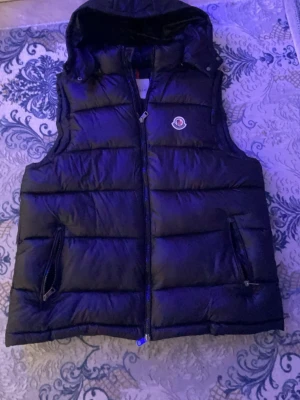Svart dunväst från Moncler - Snygg svart dunväst från Moncler med huva och dragkedja framtill. Västen har två fickor med dragkedja och det klassiska Moncler-logotypen på bröstet. Perfekt för lager på lager och streetwear-vibe.