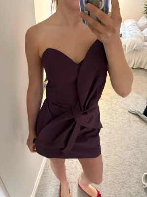 Mörklila axelbandslös kort klänning - Säljer en mörklila axelbandslös kort klänning med snygg omlottdetalj och knyt i midjan. Klänningen är helt ny med prislappen kvar😍