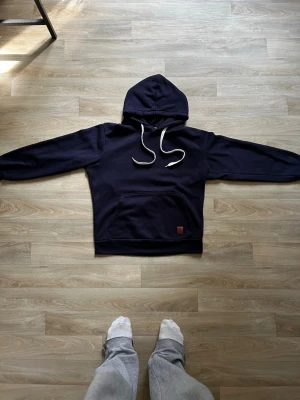 Mörkblå hoodie med vita snören - En stilren mörkblå hoodie med vita dragsnören och klassisk känguruficka framtill. Hoodien har en enkel design utan tryck och en liten brun läderlapp detalj på fickan. Perfekt för dig som gillar en clean och avslappnad look.