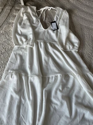 Vit känning med knyte baktill  - Oanvänd!!Luftig vit klänning  med knyte vid nacke. Klänningen har v-ringning, långa ärmar med resår vid ärmslut och ett lätt strukturerat tyg. Perfekt för varma dagar och somriga tillfällen. (Klänningen går med ungefär till knäna på mig)💕