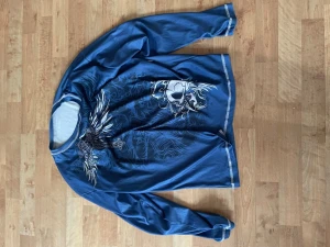 Blå stickad tröja med dödskalletryck - Cool blå tröja med stora tryck av dödskalle, vingar och kors både fram och bak. Tröjan har långa ärmar och vita detaljer längs muddar och nederkant. Perfekt för dig som gillar edgy och unika plagg.