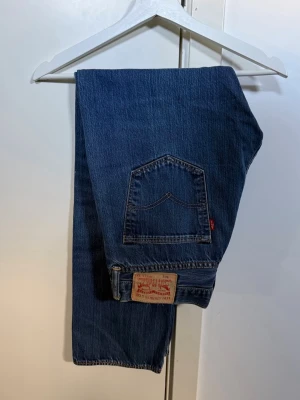 Blå Levi's 501  jeans - Klassiska blå Levi's jeans med rak passform och fem fickor. Jeansen har en ikonisk läderpatch bak och röd Levi's-tag på bakfickan. Storlek 33/32
