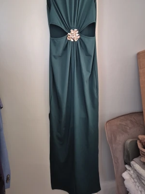 Mörkgrön långklänning med smycke - Elegant mörkgrön långklänning med dekorativt smycke i midjan och snygga utskärningar. Klänningen har en slits på sidan och är tillverkad i ett mjukt, följsamt material som ger en lyxig känsla. Perfekt för dig som vill sticka ut med en unik detalj.