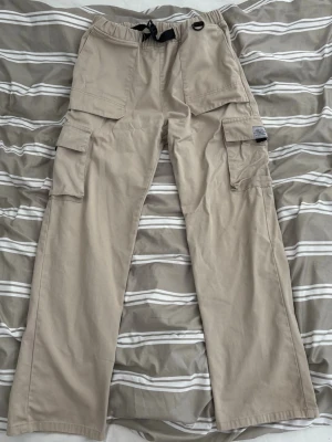 Beige cargobyxor med fickor - Snygga beige cargobyxor med flera stora fickor på sidorna och framtill. Byxorna har elastisk midja med dragsko och raka ben. Tillverkade i ett mjukt och slitstarkt material, perfekta för en avslappnad streetwear-look.