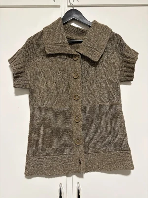 Brun stickad kofta med kort ärm - Säljer en brunmelerad stickad kofta med korta ärmar och stora träknappar framtill. Koftan har bred krage och ribbstickade detaljer vid ärmslut och nederkant. Perfekt för lager på lager och en avslappnad stil.