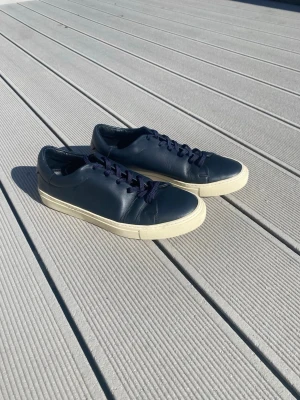 Mörkblå sneakers i läder - Snygga mörkblå sneakers av Sandro Paris i slätt läder med matchande mörkblå skosnören och vit sula. Klassisk låg modell som passar till det mesta och ger en clean look. Perfekta för dig som gillar minimalistisk stil.