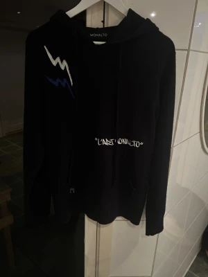 Merinoull hoodie från Monalto - Svart hoodie från Monalto med broderad vit och blå blixtdetalj på bröstet och texten 'LAST MONALTO' framtill. På ryggen står det 'MADE IN FRANCE' i vit text. Hoodien har huva och dragsko. Perfekt för en streetwear-look.