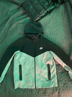 Nike tvåfärgad hoodie med dragkedja - Snygg hoodie från Nike i svart och ljusgrått med dragkedja framtill och två fickor med svarta dragkedjor. Klassisk Nike-logga på bröstet och huva för extra stilpoäng. Perfekt för dig som gillar sportig och avslappnad look. Storlek L 