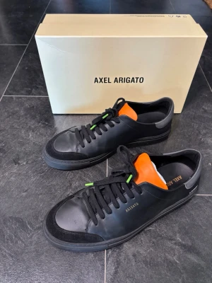 Axel Arigato sneakers - Säljer ett par Axel Arigato sneakers i svart. Riktigt snygga med orange detalj, sticker ut lagom. Använda ett fåtal gånger, i näst intill toppen skick. Kommer med original box. Hör av dig vid intresse 🙌