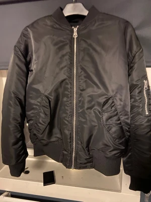 Svart bomberjacka med dragkedja - Klassisk svart bomberjacka med silvrig dragkedja framtill och ribbade muddar vid ärmslut och midja. Jackan har två snedställda fickor fram och en ficka med dragkedja på ena ärmen. Tillverkad i glansigt syntetmaterial för en clean och stilren look.