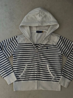 Randig hoodie från Polo Ralph Lauren - Snygg vit hoodie med marinblå horisontella ränder från Polo Ralph Lauren. Dragkedja framtill, två fickor och klassisk huva. Liten broderad logga på bröstet. Tillverkad i mjukt bomullsmaterial som känns skönt mot huden. Perfekt för en avslappnad och stilren look. Hoodien är i 9/10 i skick. Tröjan är i storlek M men passar även S!