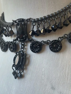 Svart statement choker med pärlor - Unikt svart choker-halsband med pärlor och dekorativa detaljer. Halsbandet har en gotisk vibe med svarta pärlor och hängande dekorationer i plast. Perfekt för att lyfta en outfit med en edgy touch.