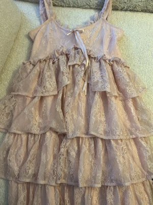 Ljusrosa volangklänning i spets - Romantisk ljusrosa klänning med flera lager volanger och spetsdetaljer. Klänningen har smala axelband, omlottkänsla framtill med liten rosett och är tillverkad i ett tunt, luftigt material. Perfekt för sommarens alla tillfällen.