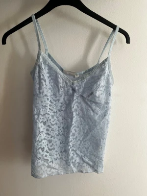 Ljusblå spetslinnetopp från Kappahl - Säljer en ljusblå linnetopp från Kappahl med tunna axelband och v-ringning. Toppen är täckt av ett fint spetsmönster som ger en romantisk vibe. Perfekt att styla med jeans eller kjol för en trendig look.