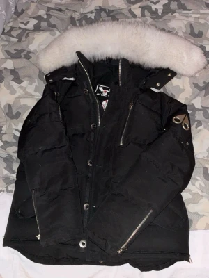 Svart  Moose Knuckles - Snygg svart dunjacka från Moose Knuckles med stor vit pälskantad huva, dragkedjor vid ärmslut och bröstficka samt metallogo på ärmen. Jackan har en klassisk puffer-modell och är perfekt för kalla vinterdagar.