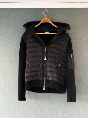 Moncler cardigan-svart - Svart cardigan från Moncler med quiltad front och huva. Jackan har ribbstickade ärmar och sidor, dragkedja framtill. Hängande moncler loggan har slitage men den kan tas av om man hellre vill det. Den är M men passar också S väl.